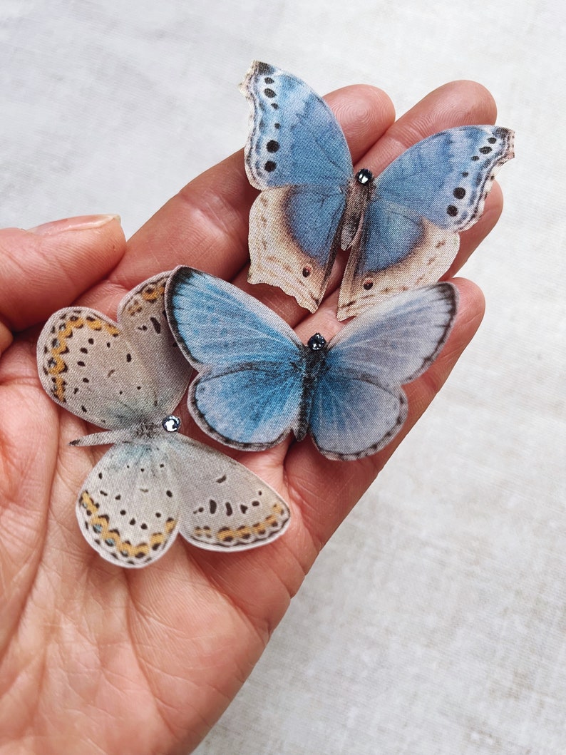A hand holds three palest blue silk butterflies. Each little butterfly has a tiny sparkling crystal on its head. these silk butterflies are slipped onto bobby pins and are the perfect wedding accessory.