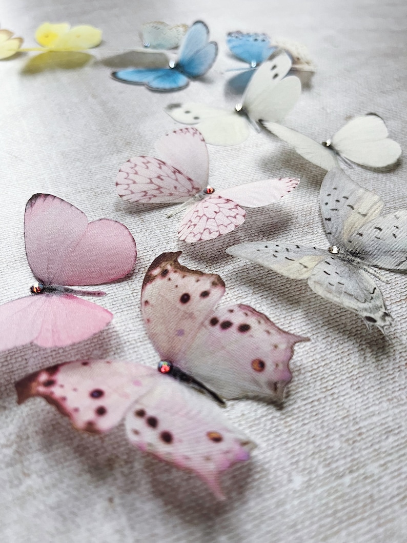 Perfectly hand cut from fine silk a selection of pastel butterflies sit on a pale linen cloth. The silk butterflies are pale pink, white, pale blue and pale yellow in colour. each one has a tiny sparkling crystal on its head.