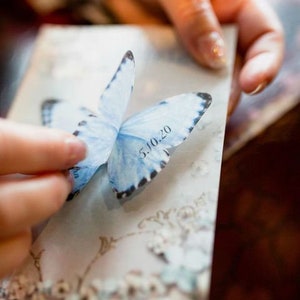 Hands hold a postcard with a something blue silk butterfly, the fingers are lifting the wing to show the wedding date custom printed on the lower wing