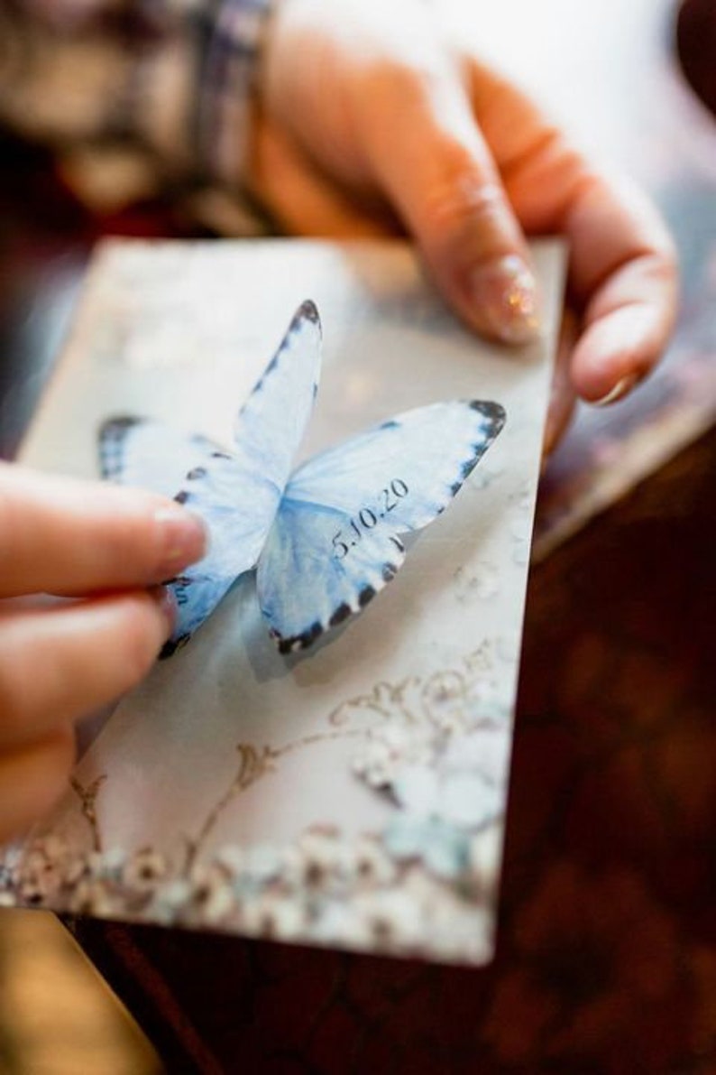 Hands hold a postcard with a something blue silk butterfly, the fingers are lifting the wing to show the wedding date custom printed on the lower wing