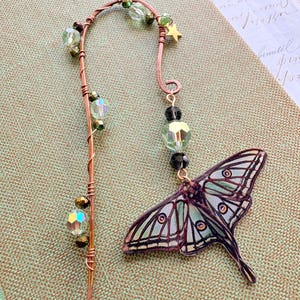 May include: A copper metal bookmark with a decorative design. The bookmark features a moth pendant with green and black accents, along with green and clear beads and a small gold star. The bookmark is placed on a green surface.