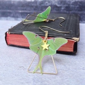 A pair of brass triangle earrings with a green silk luna moth design. Each earring features a small gold star charm.