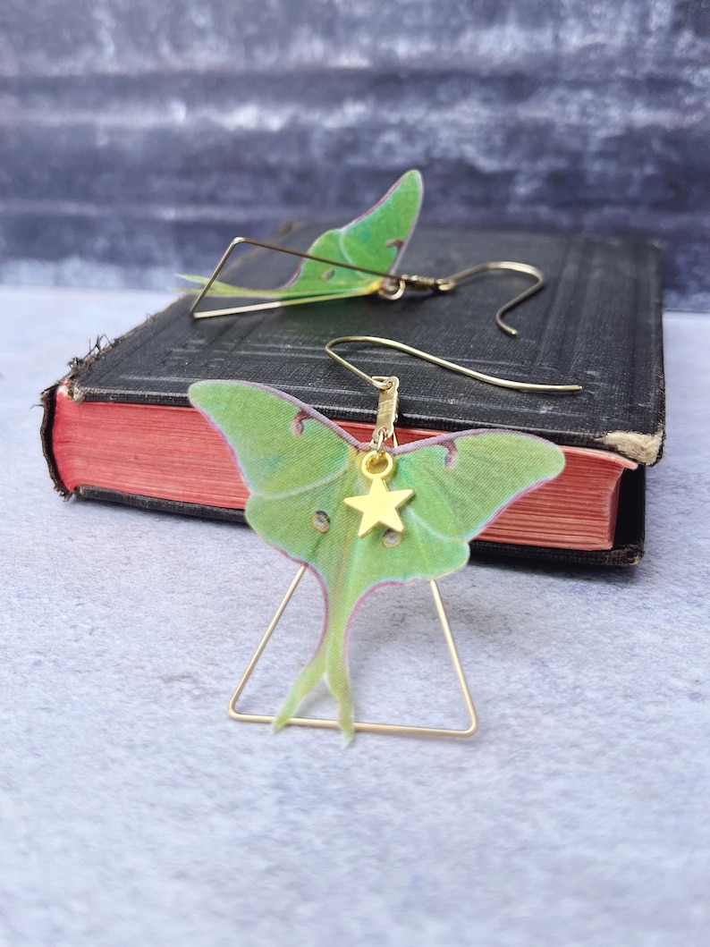 A pair of brass triangle earrings with a green silk luna moth design. Each earring features a small gold star charm.