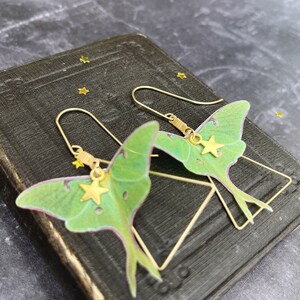 Two green and brass earrings shaped like luna moths with gold star charms hanging from them.