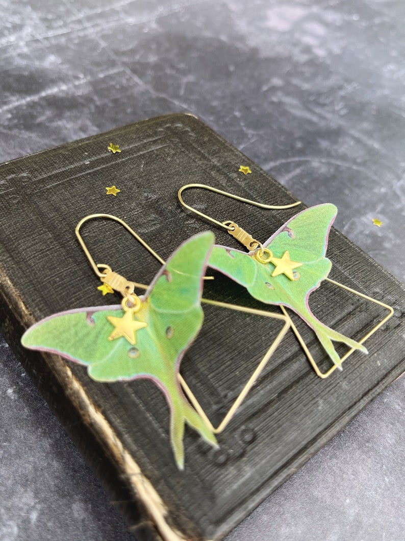 Two green and brass earrings shaped like luna moths with gold star charms hanging from them.