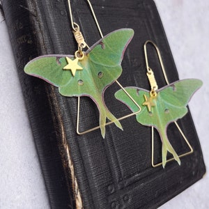 Two brass triangle earrings with green silk moth wings and gold star charms sit on a vintage black prayer book