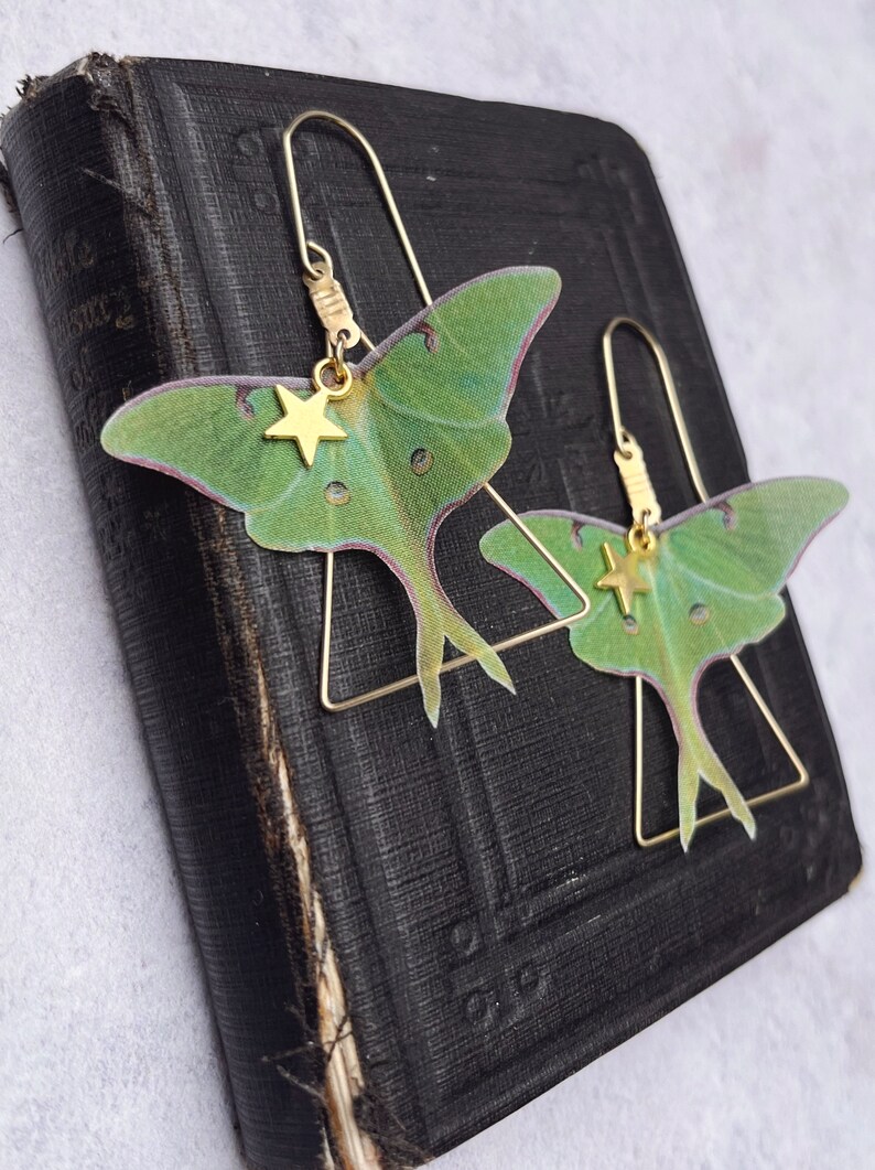 Two brass triangle earrings with green silk moth wings and gold star charms sit on a vintage black prayer book