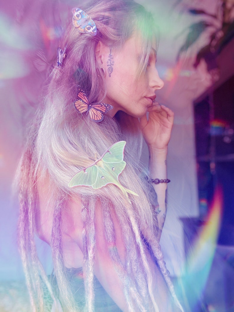 An image of a lady with long blond dreads wears a selection of silk butterfly hair clips and a large green luna moth hair clip. the image is slightly blurred with rainbow bokeh lights