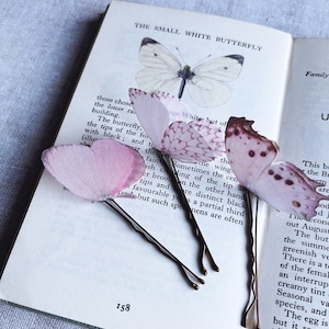 Perfect wedding hair accessories, 3 palest pink butterfly hair pins sit on an opened vintage butterfly book. These butterflies have their wings folded and are super realistic, it looks like real butterflies have landed in your hair!