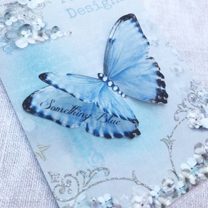 A pale blue silk butterfly hair clip with the words something blue on the lower wings. It is sitting on a pretty vintage style glossy gift card