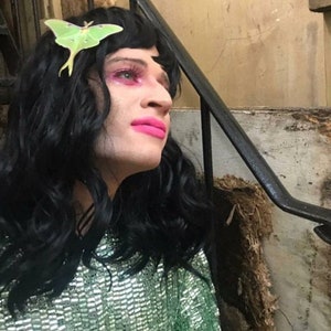 A customer review image of a man with long black hair and bright pink lips and eye shadow. he wears a green sequinned top and a luna moth hair clip in his hair.