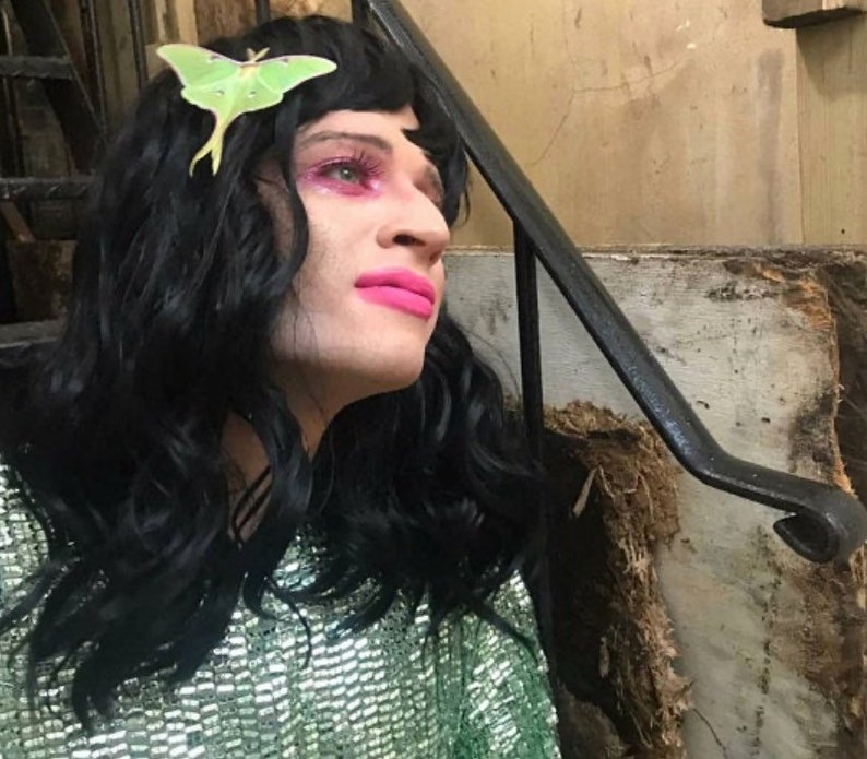 A customer review image of a man with long black hair and bright pink lips and eye shadow. he wears a green sequinned top and a luna moth hair clip in his hair.