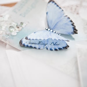 On a pretty vintage style glossy postcard sits a pleats blue silk butterfly with a pale blue crystal body. The butterfly has a double layer wing and on the bottom wing are the words something blue.