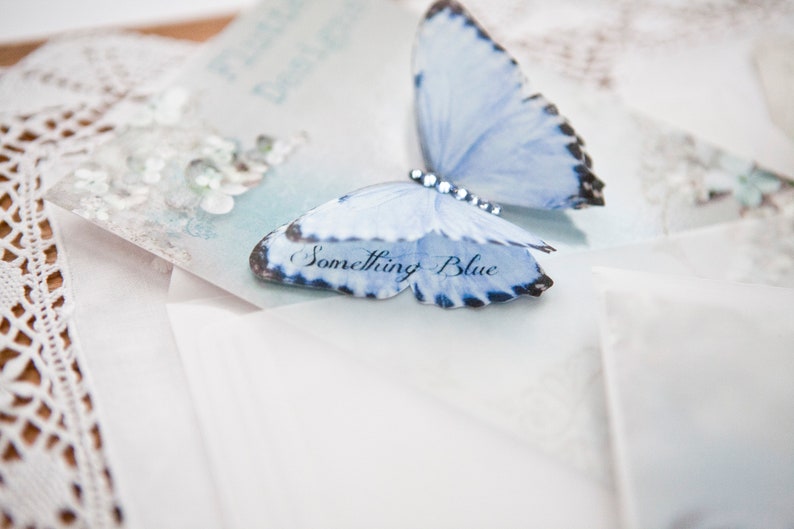 On a pretty vintage style glossy postcard sits a pleats blue silk butterfly with a pale blue crystal body. The butterfly has a double layer wing and on the bottom wing are the words something blue.