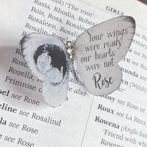 以下が含まれることがあります： 白地にグレーと白の模様の入った羽根を持つ白い蝶。蝶は「あなたの翼は準備万端でしたが、私たちの心は準備万端ではありませんでした。ローズ」というテキストが書かれたページの上にいます。