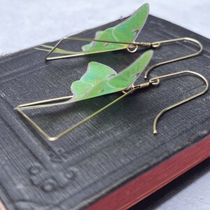 Two brass triangle shaped earrings with green moth wings attached. The wings are translucent and have a light green color.