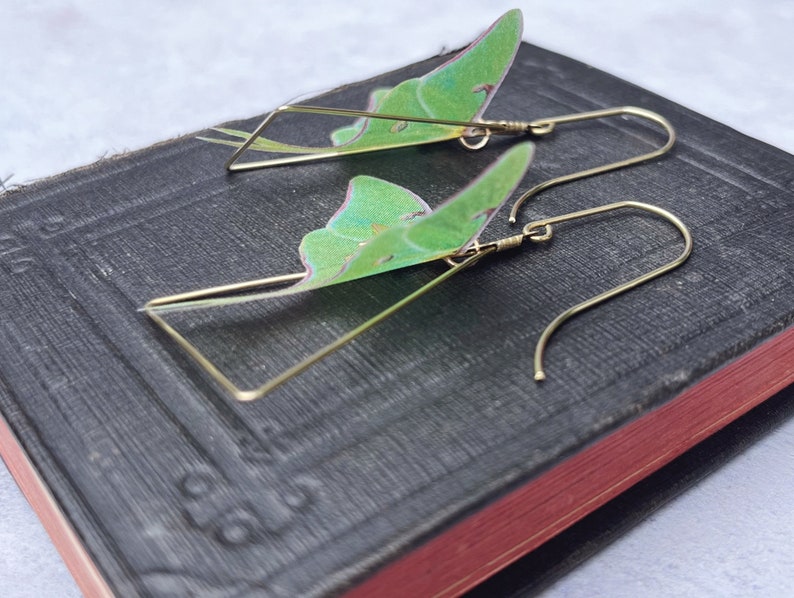 Two brass triangle shaped earrings with green moth wings attached. The wings are translucent and have a light green color.