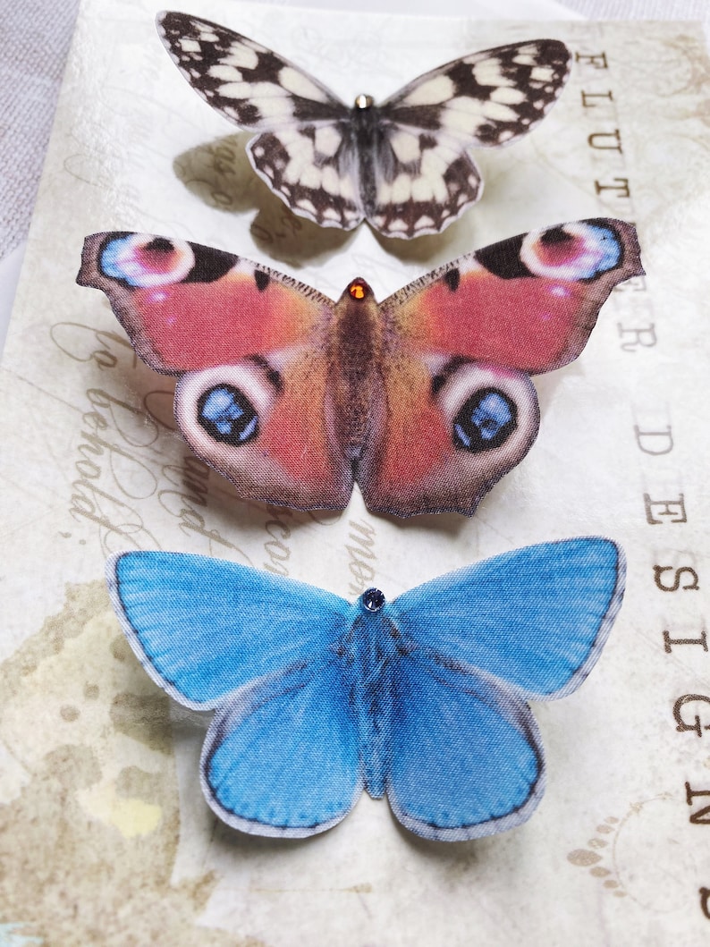A marbled white butterfly, a peacock butterfly and a blue adonis butterfly sit on a glossy presentation postcard. The butterflies are super realistic silk hair clips