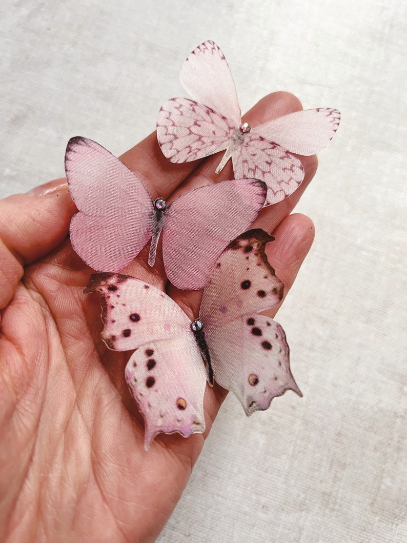 A hand holds a set of 3 pale pink silk butterflies in different shapes. Each one has been carefully hand cut from fine silk and has a tiny sparkling crystal on its head. Pretty summer wedding accessories