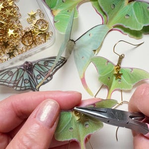 A maker is creating a pair of green luna moth earrings, they are using pliers to attach the small gold Star charms