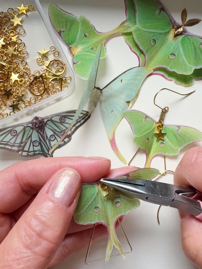 A maker is creating a pair of green luna moth earrings, they are using pliers to attach the small gold Star charms