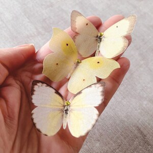 A hand holds 3 pale yellow silk butterflies with crystal details. each butterfly is hand cut from fine silk that has been gently stiffened so they hold their shape and do not fray. They are super realistic!