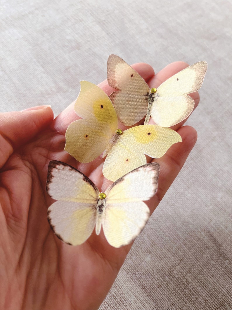 A hand holds 3 pale yellow silk butterflies with crystal details. each butterfly is hand cut from fine silk that has been gently stiffened so they hold their shape and do not fray. They are super realistic!