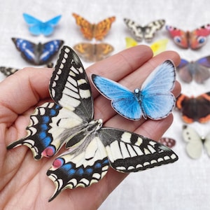 A  hand holds a large swallowtail butterfly and a small holly blue butterfly. The butterflies are hand cut from silk and have little sparkling crystals on their heads
