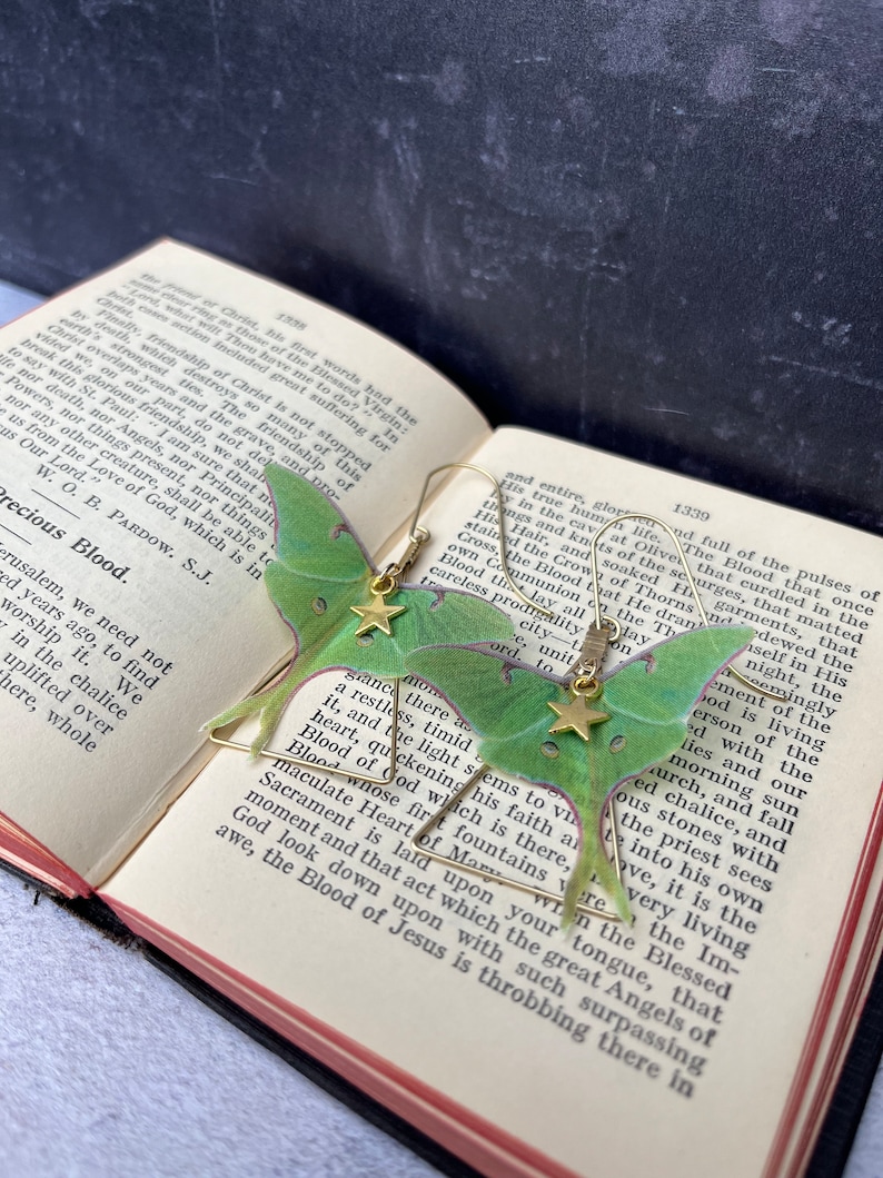 Two brass earrings with a green moth design. The moths have gold stars on their wings. The earrings are hanging from a book with text on the pages. The text on the pages reads: 'Precious Blood.'