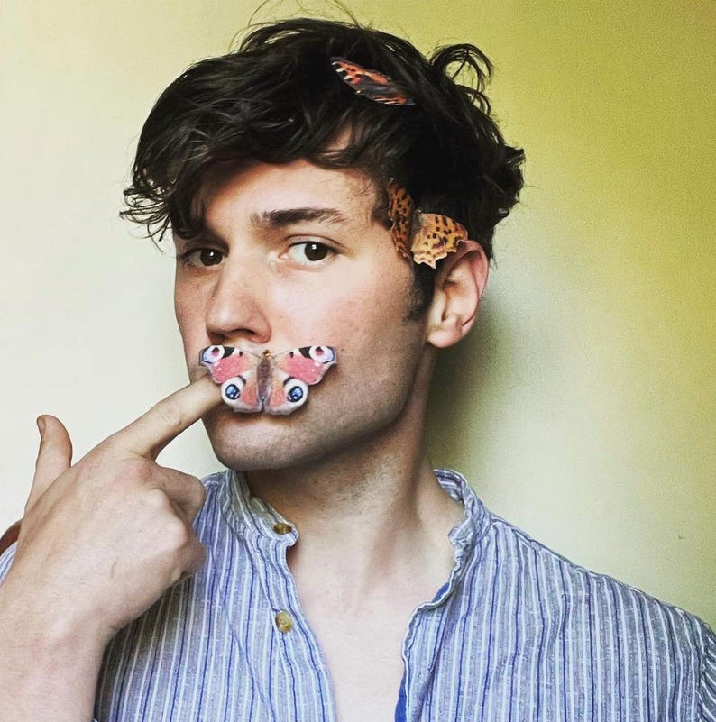 a man in a blue pin stripe shirt unbuttoned at the neck wears two silk butterfly hair clips in his hair and a peacock butterfly on his finger that he has on his lip