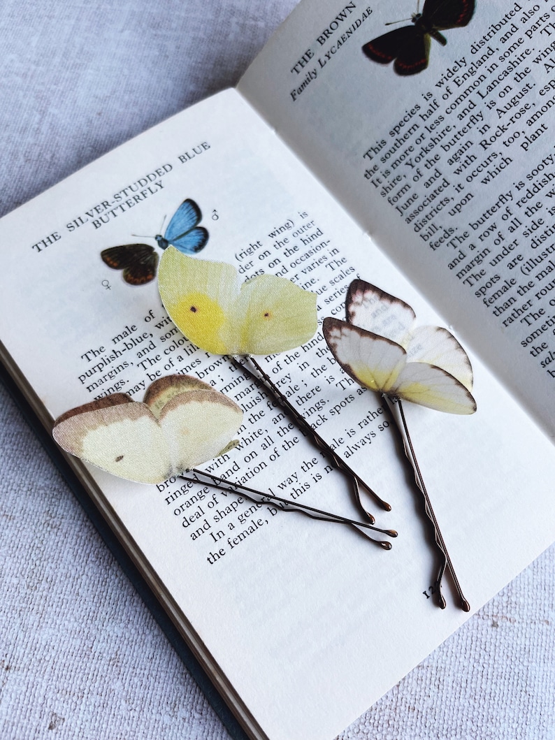 3 silk butterfly wedding hair pins sit on an open vintage butterfly book. The butterflies are palest yellow and have their wings folded like real butterflies