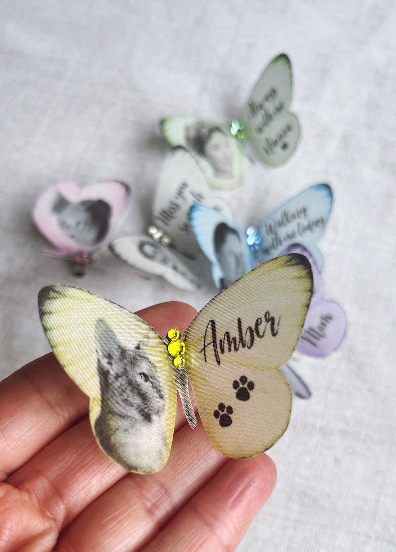 A little pale yellow silk butterfly sits on finger tips. On the butterfly are an image of a cat and the name amber. there are also paw prints and co ordinating coloured crystals