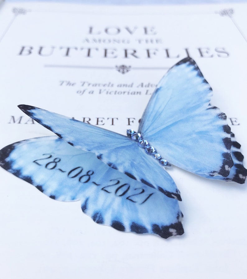 A blue silk butterfly with the date 28th August 2021 hidden under the wing. the body is covered with pale blue crystals.