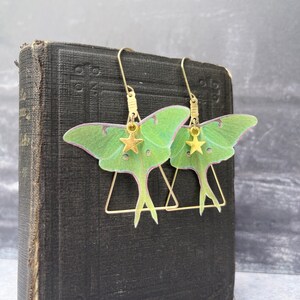 May include: A pair of gold earrings featuring green and pink moth wings with gold star charms hanging from the bottom. The earrings are hanging from a book.