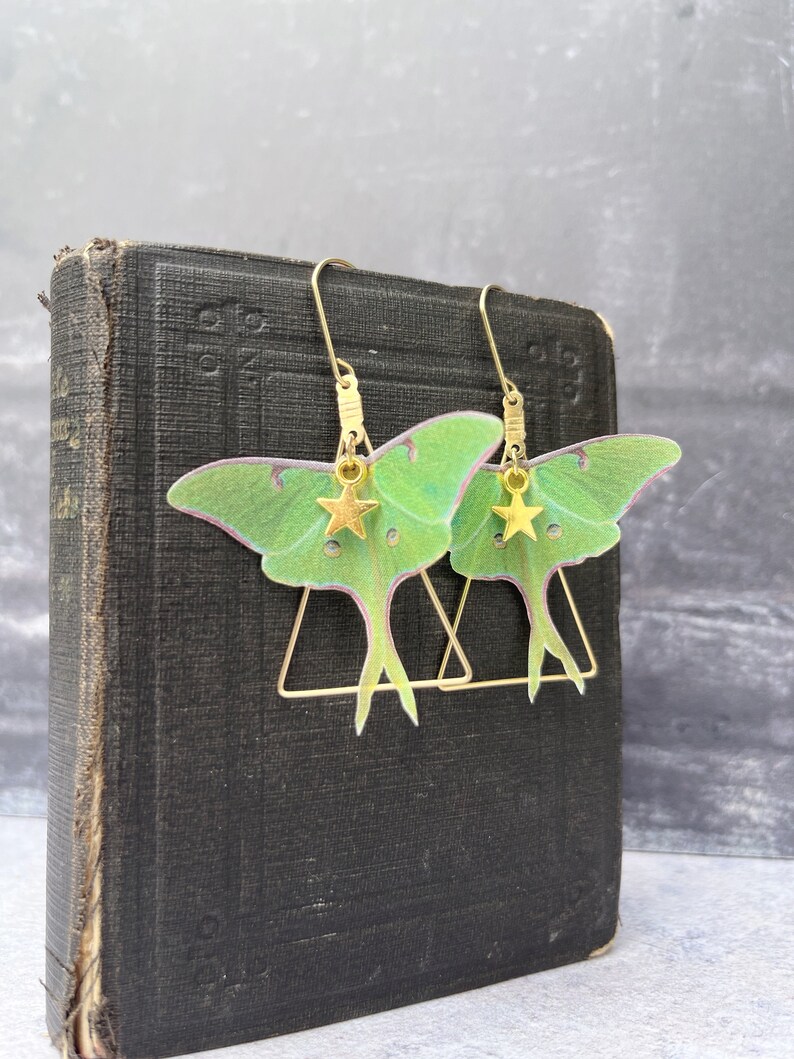 May include: A pair of gold earrings featuring green and pink moth wings with gold star charms hanging from the bottom. The earrings are hanging from a book.
