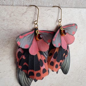 May include: A pair of butterfly wing earrings. The earrings have gold-coloured hooks and small red gemstones. The wings are a mix of black, orange, pink, and grey, with a detailed pattern. The earrings are designed to resemble butterfly wings.