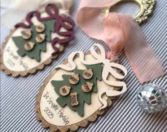 May include: Two oval wooden ornaments featuring a scalloped edge, a green Christmas tree cutout, and small wooden tags engraved with names. One ornament has a cream ribbon and the other a dusty pink ribbon. Text reads The Snyder Family 2025.