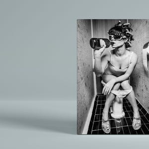 Puede incluir: Foto en blanco y negro de una mujer sentada en un inodoro en un ba&ntilde;o, bebiendo de una botella. Lleva gafas de sol y un vestido. El inodoro es blanco y las paredes del ba&ntilde;o est&aacute;n enlosadas.