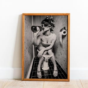Puede incluir: Fotograf&iacute;a en blanco y negro de una mujer sentada en un inodoro en un ba&ntilde;o, bebiendo de una botella. Lleva gafas de sol y tiene el pelo en rulos. La foto est&aacute; enmarcada en un marco de madera.