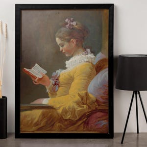 Puede incluir: Una pintura de una mujer con un vestido amarillo leyendo un libro. Está sentada en una silla con un cojín azul. La pintura está en un marco negro.