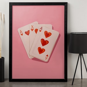 Puede incluir: Una impresión enmarcada con tres cartas de juego con corazones rojos sobre un fondo rosa. Las cartas son el As de Corazones y una tercera carta con un corazón. El marco es negro.