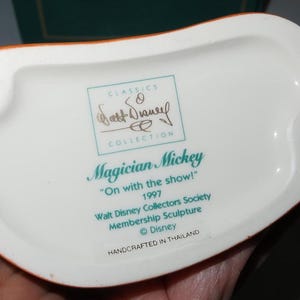 May include: Underside of a white ceramic figurine base with an orange rim. Text includes "Magician Mickey", "On with the show!", and "Walt Disney Collectors Society". The base has two holes and is marked "Handcrafted in Thailand".