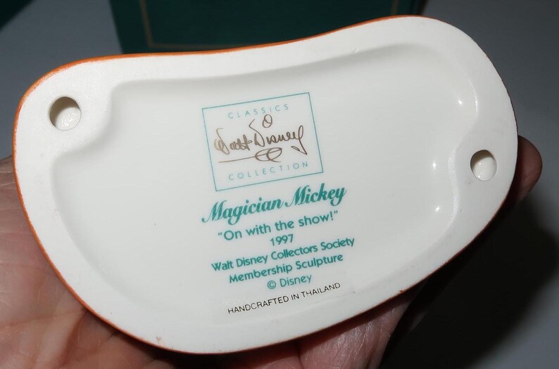 May include: Underside of a white ceramic figurine base with an orange rim. Text includes "Magician Mickey", "On with the show!", and "Walt Disney Collectors Society". The base has two holes and is marked "Handcrafted in Thailand".