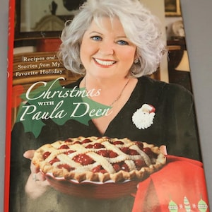 May include: A cookbook titled "Christmas with Paula Deen" featuring a photo of Paula Deen holding a cherry pie. The book cover is red, green, and white with a festive Christmas theme.
