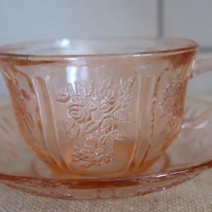 May include: A pink glass teacup and saucer set with a floral design. The cup and saucer are both decorated with a delicate pattern of roses.