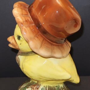May include: A ceramic figurine of a yellow duck wearing a brown hat. The duck is perched on a base with green and brown painted details. The hat has a wide brim and a rounded top. The duck has a small beak and a dark eye.
