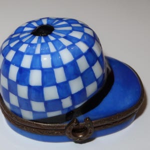 May include: A blue and white checkered hat-shaped trinket box. The hat has a blue brim and a brown metal band with a horseshoe clasp. The top of the hat is a blue and white checkered pattern.
