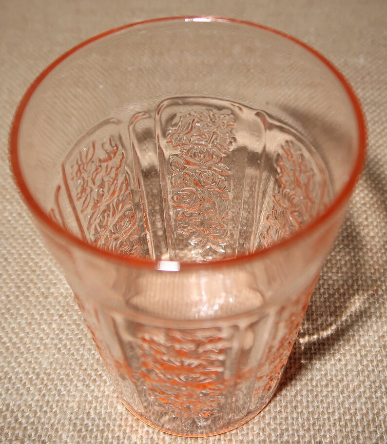 May include: A clear pink glass tumbler with a floral pattern. The glass has a slightly rippled texture and a rounded bottom.