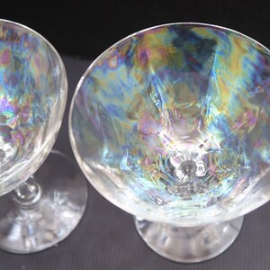 May include: Two clear iridescent stemmed glasses. The bowls of the glasses reflect a rainbow of colors, including blue, green, and yellow. The stems and bases are clear and simple, with a small floral design at the base of the bowl.