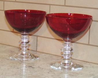 May include: Two ruby red stemmed glasses with a wide bowl and a clear, decorative stem. The stem has a stacked ring design. The glasses sit on a clear, round base. The glasses are on a light-colored surface.